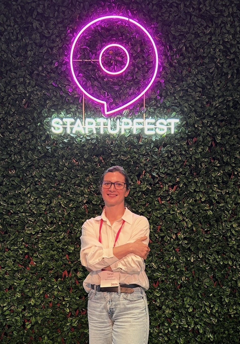 A person standing beneath an illuminated Startupfest sign against a leafy wall.